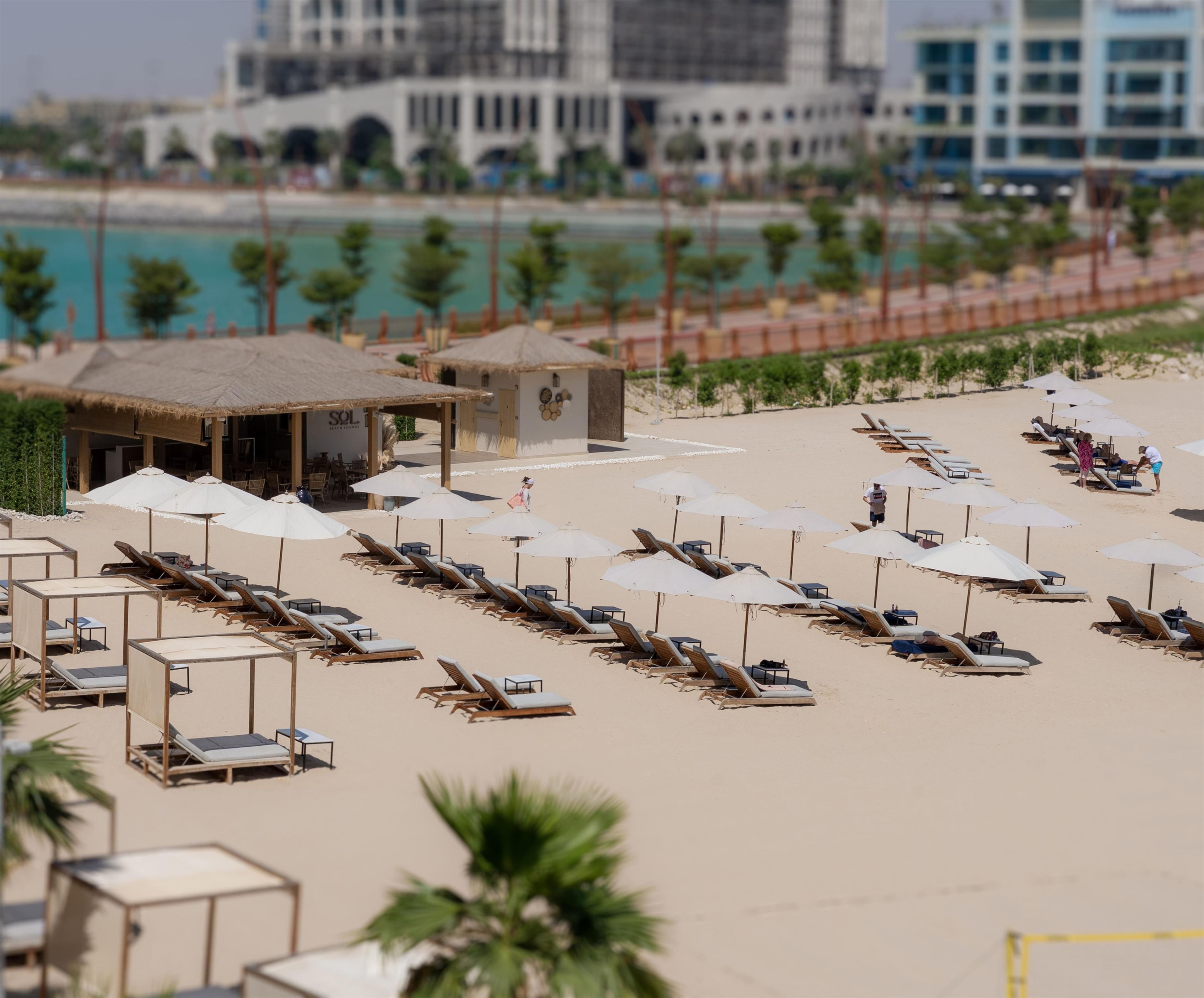 A beach with sun loungers and umbrellas, a small hut, and a building in the background.