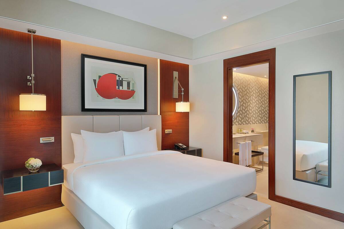 Une chambre d’hôtel moderne avec un grand lit, des lampes de chevet, une œuvre d’art encadrée et une salle de bain visible à travers une porte ouverte.