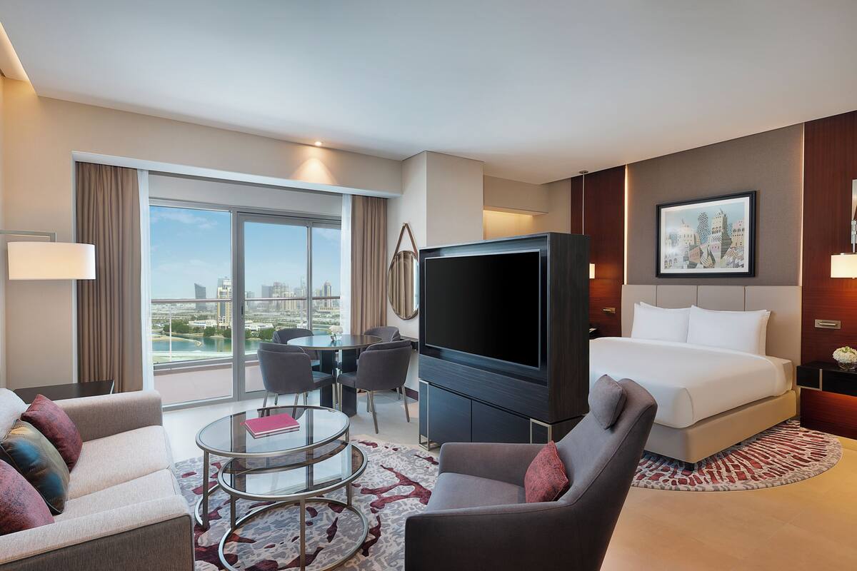 Une chambre d’hôtel moderne dotée d’un grand lit, d’un téléviseur à écran plat, d’un coin repas avec une table et des chaises, et offrant une vue sur le paysage urbain grâce à de grandes fenêtres.