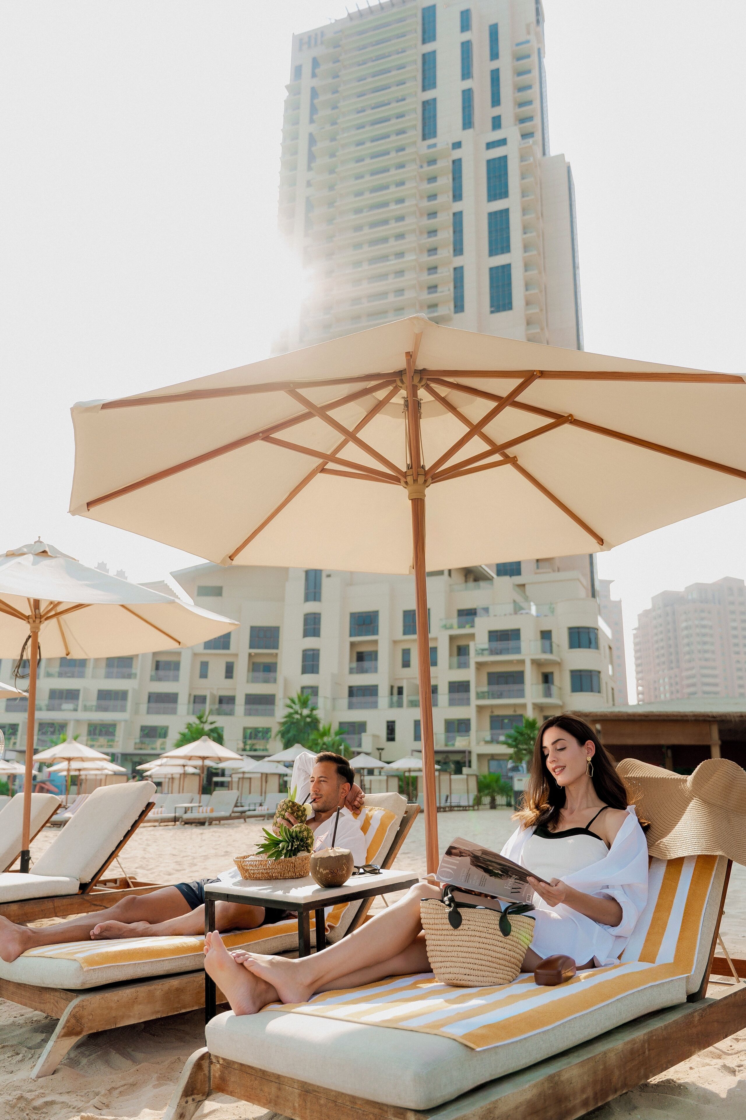 Un espace de détente aménagé sur la plage, avec des chaises longues, des parasols et un immeuble de grande hauteur en arrière-plan.
