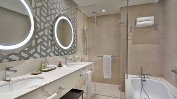 Une salle de bain moderne avec deux lavabos, une douche à l’italienne, un grand miroir avec éclairage intégré et une baignoire.