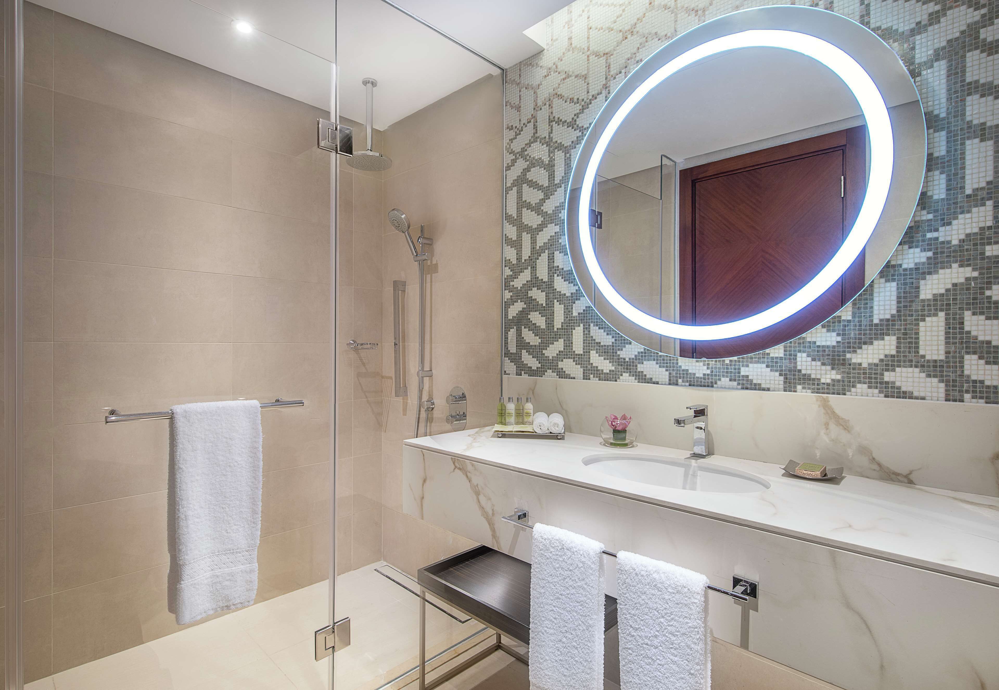 Un bagno moderno con doccia vetrata, lavabo in marmo e specchio con illuminazione circolare.