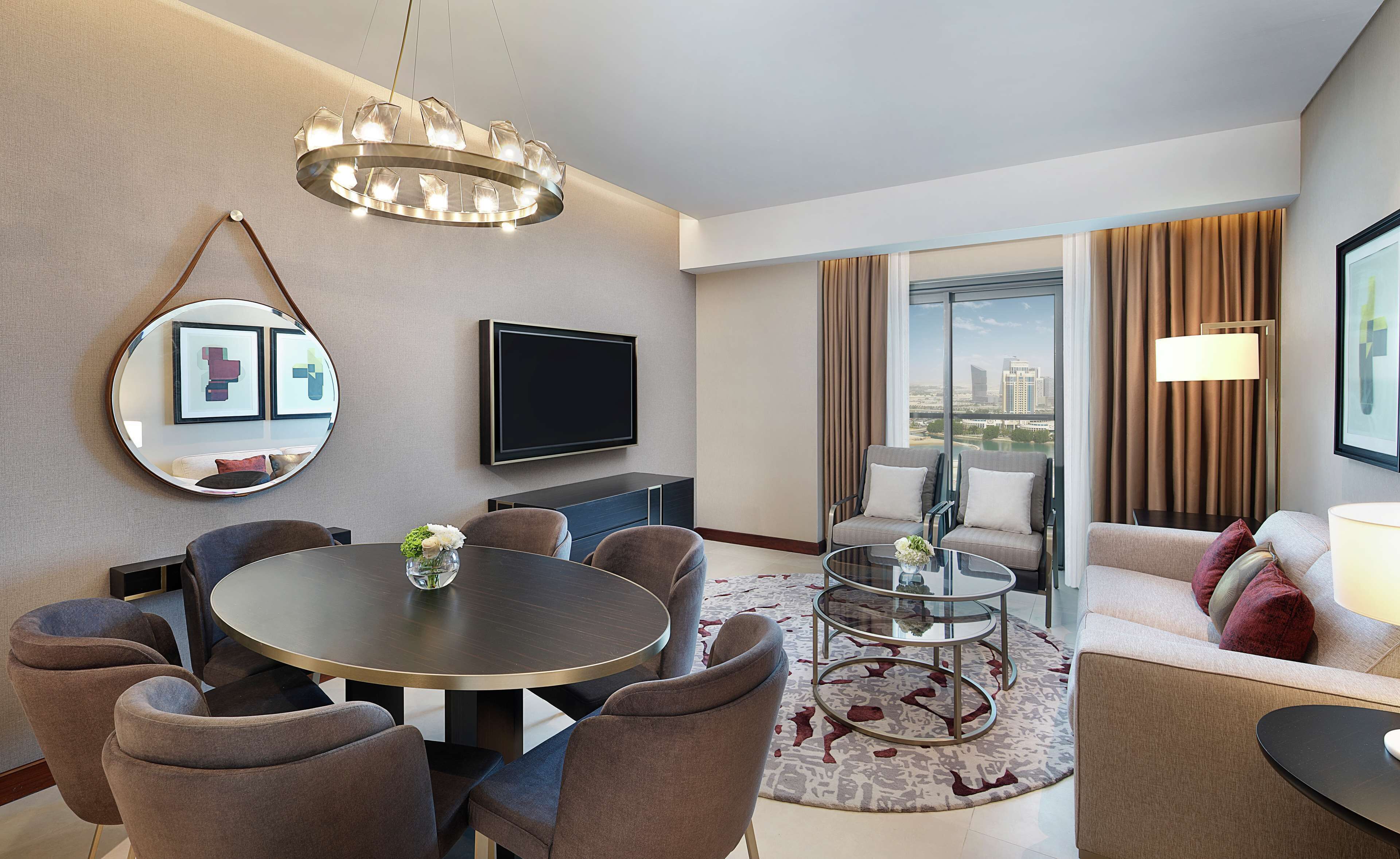 Een moderne hotelkamer met een ronde eettafel, comfortabele zitplaatsen, een grote spiegel, een flatscreen televisie en uitzicht op de stad door grote ramen.