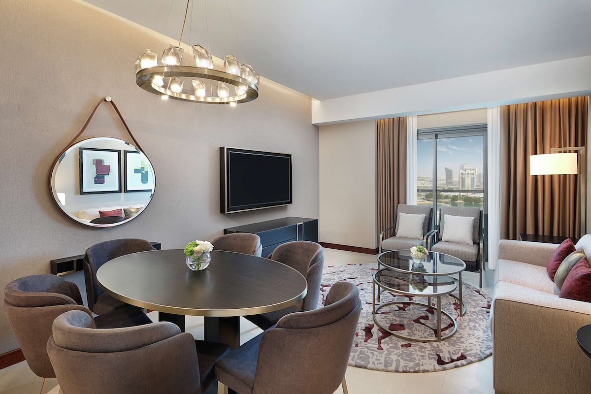 Quarto de hotel moderno com uma mesa de jantar redonda, assentos confortáveis, um espelho grande, TV de tela plana e vista da cidade através de amplas janelas.