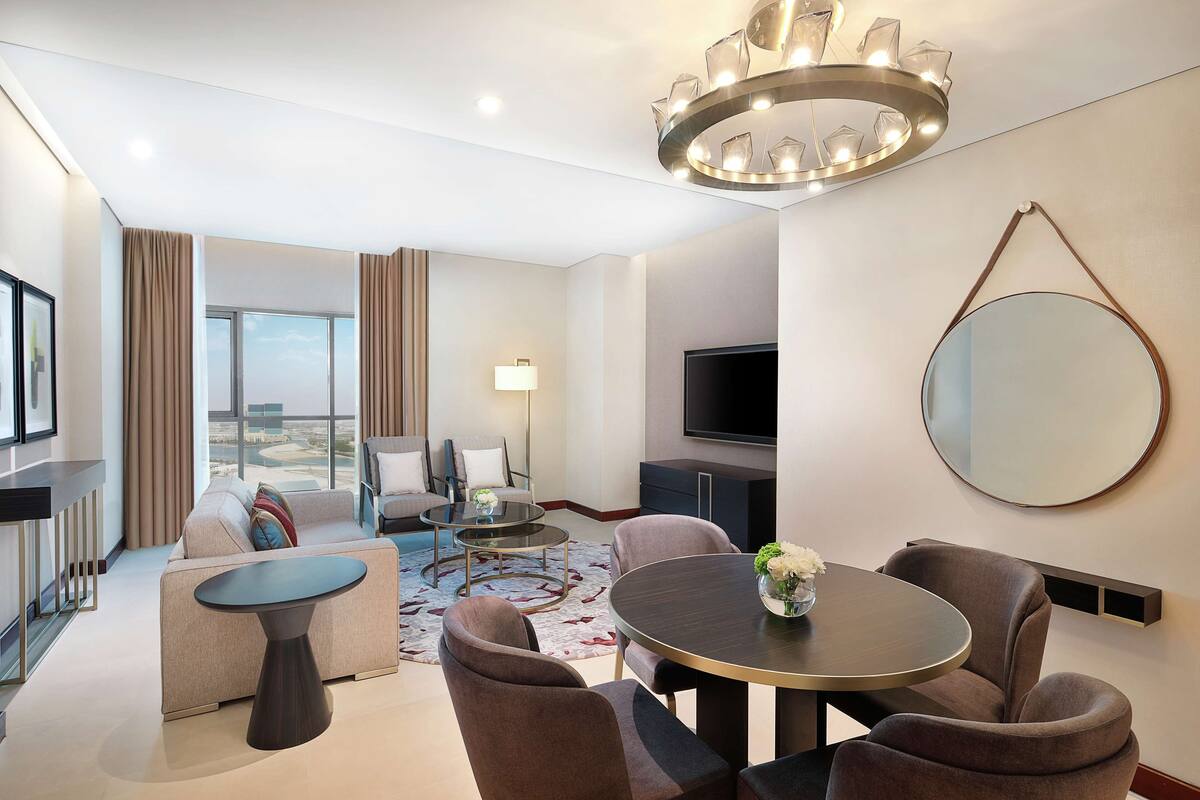 Une chambre d’hôtel moderne équipée d’un canapé, de deux fauteuils, d’une table basse, d’une table à manger, d’un téléviseur et d’un grand miroir.