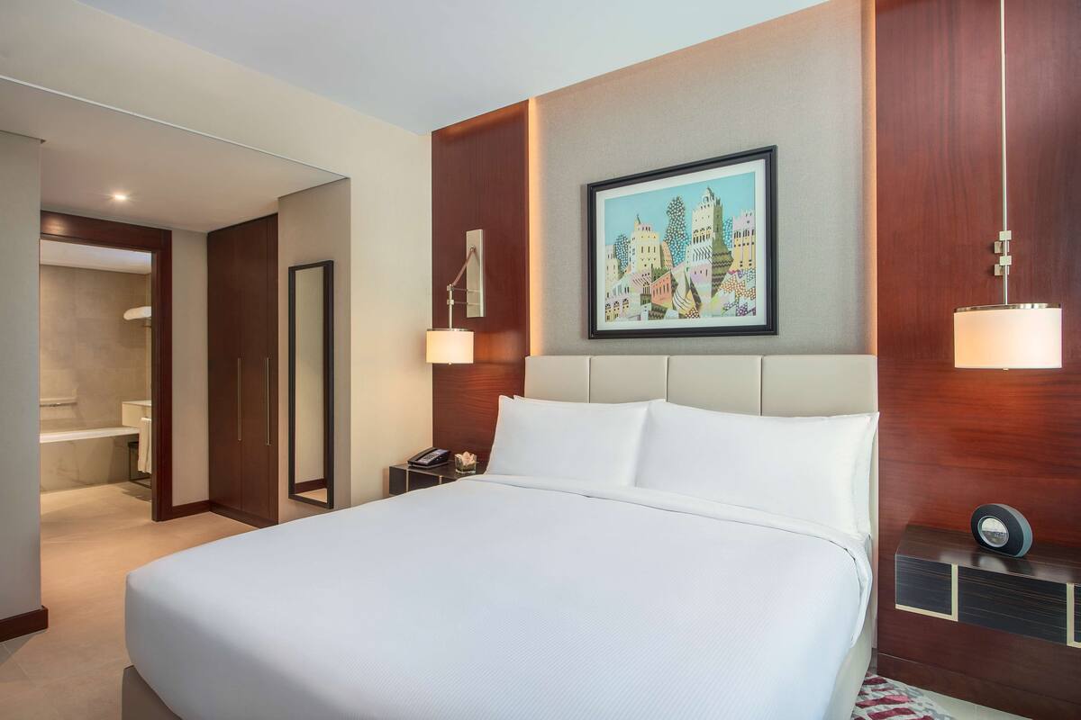 Une chambre d’hôtel avec un grand lit, une table de chevet avec une lampe, un tableau encadré au mur et une salle de bain visible à travers une porte ouverte.