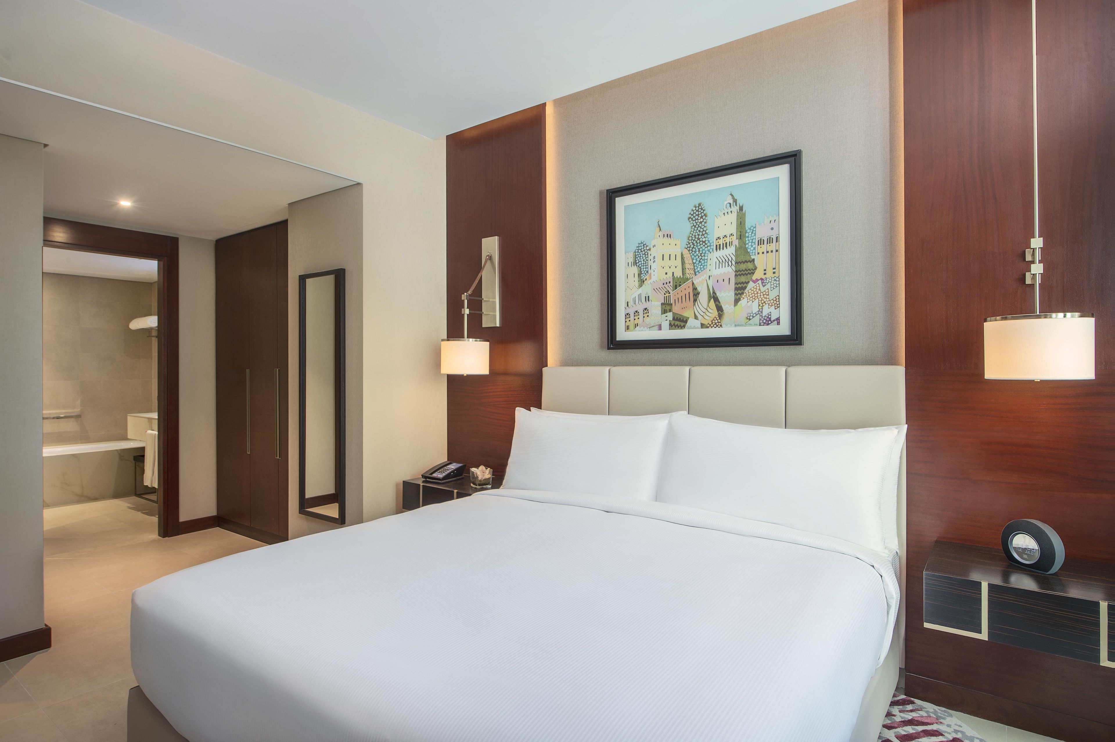 A hotel room with a large bed, a bedside table with a lamp, a painting on the wall, and a bathroom visible through an open door.