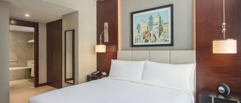 A hotel room with a large bed, a bedside table with a lamp, a painting on the wall, and a bathroom visible through an open door.