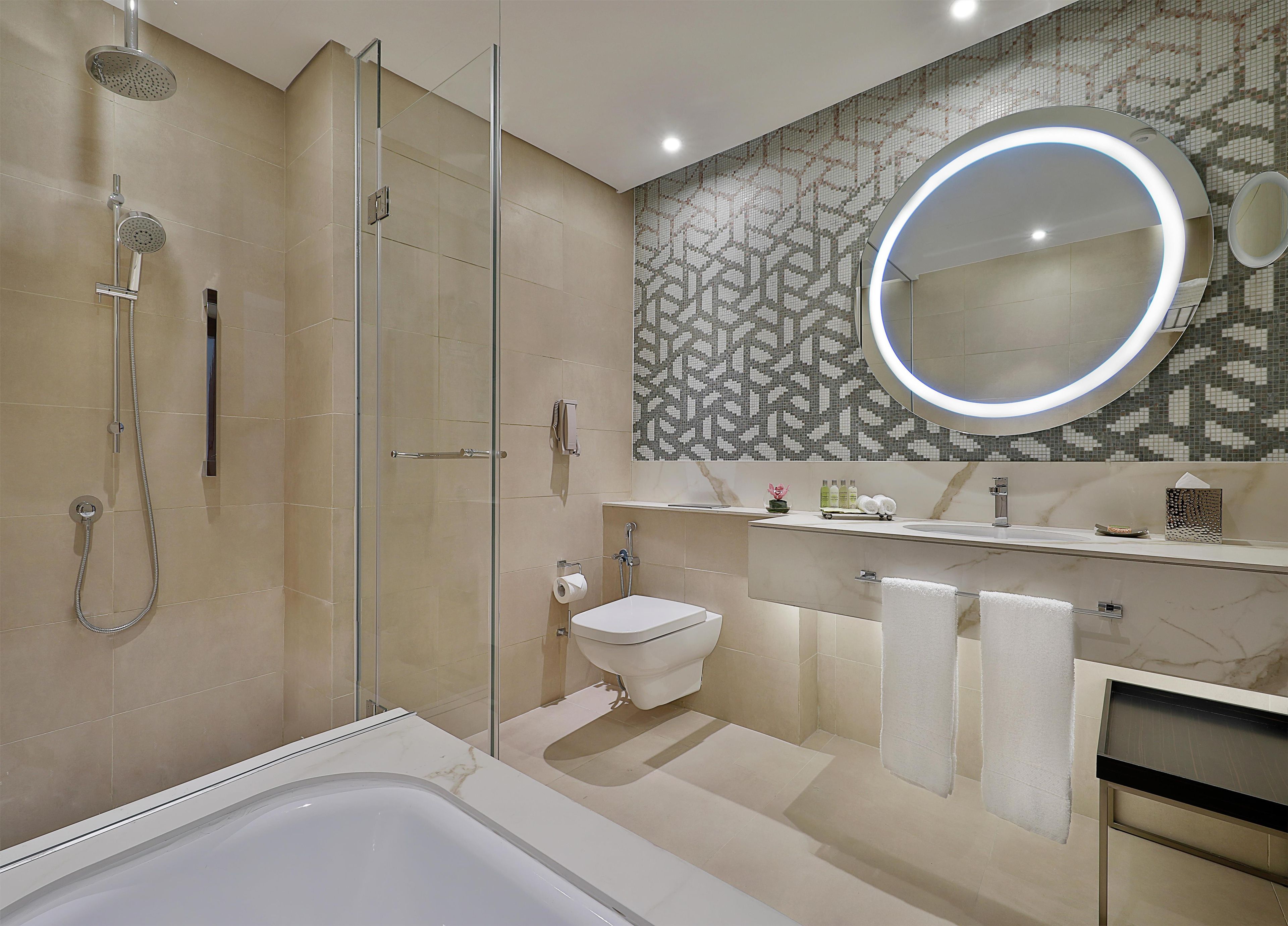 A modern bathroom with a glass-enclosed shower, a freestanding bathtub, a round mirror with integrated lighting, and a patterned backsplash.