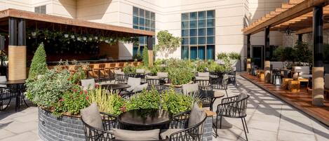Un patio al aire libre con mesas y sillas redondas, rodeado de plantas en macetas y una zona de bar con plantas colgantes.