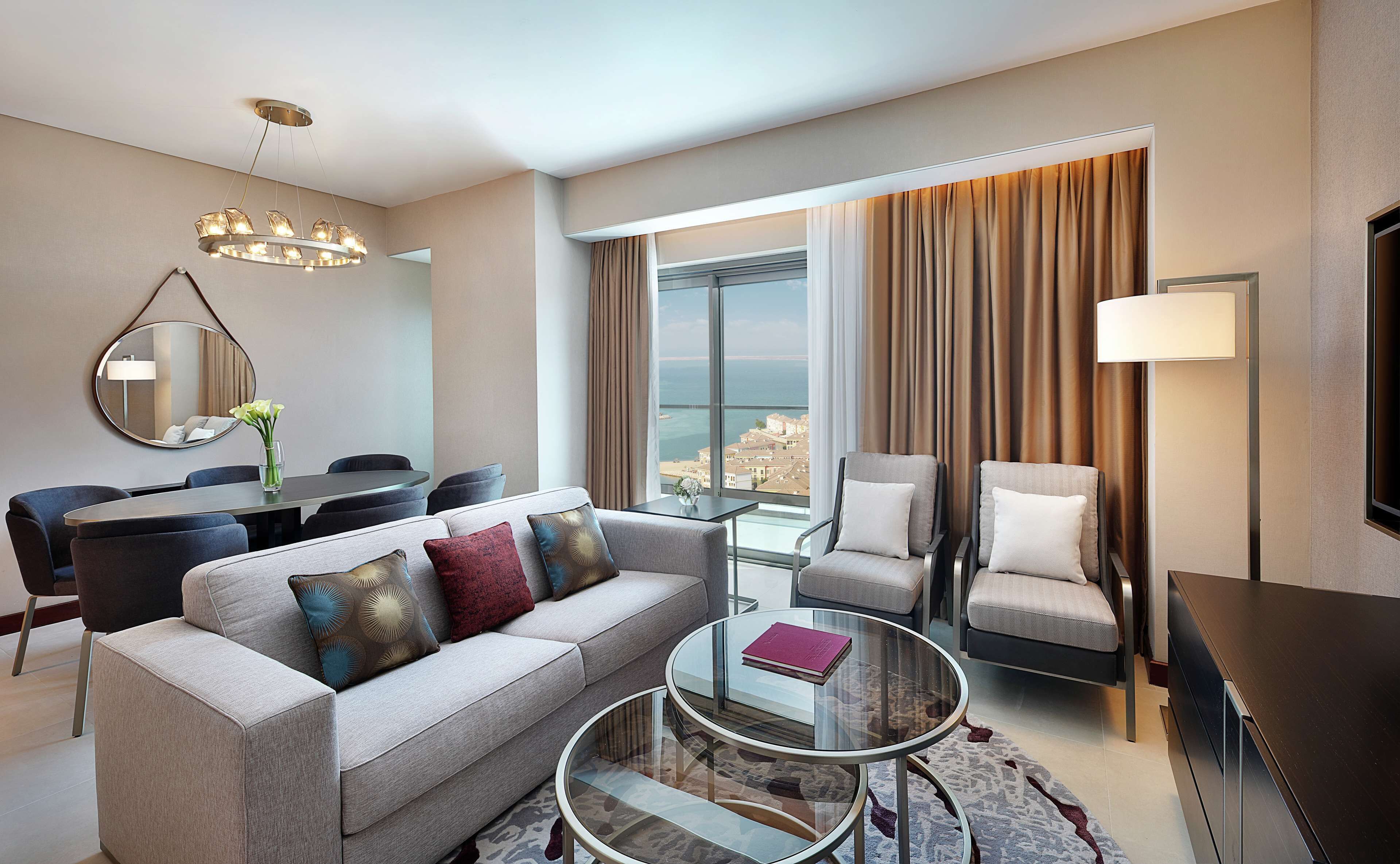 Una habitación de hotel moderna con sofá, sillones, zona de comedor y un gran ventanal con vistas a la ciudad.