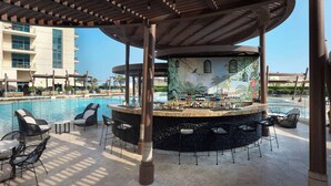 Zona de bar junto a la piscina con un mural, sillones de descanso y vistas a un edificio de hotel.