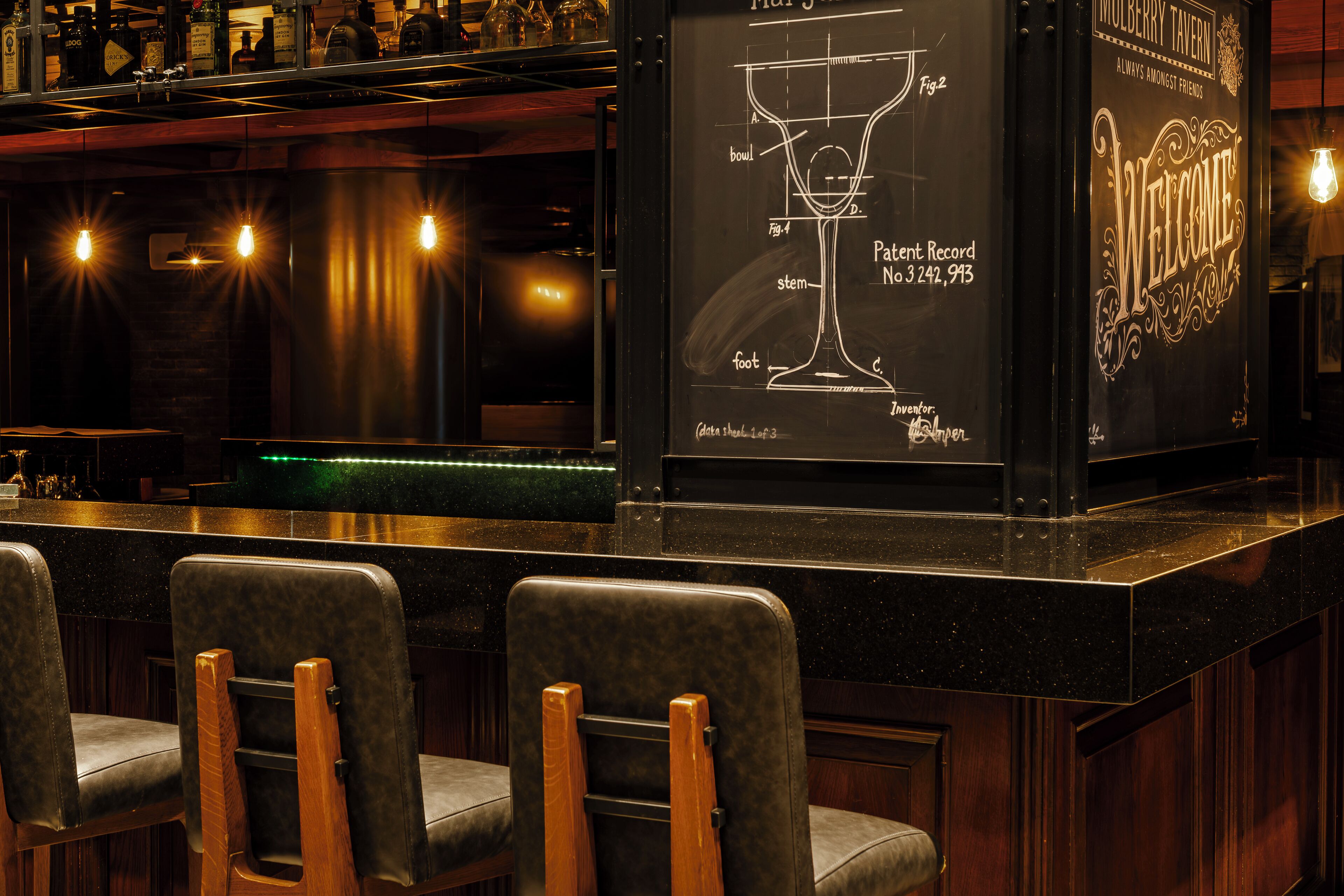 A bar area with a chalkboard displaying a drink recipe diagram, bar stools, and a 'Welcome' sign.