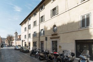 Ruas de calçada com scooters e carros estacionados, um edifício com janela redonda e uma torre ao fundo.