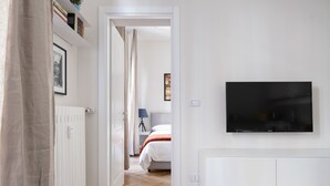 A modern hotel room with a flat-screen TV mounted on the wall, a bed with white bedding, a bedside table with a lamp, and a door leading to another room.
