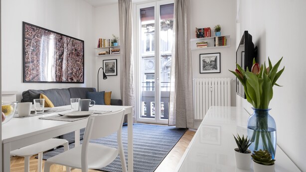 A modern living room with a white sofa, a dining table with chairs, a large abstract painting, a bookshelf, and a view through a glass door to an outdoor balcony.