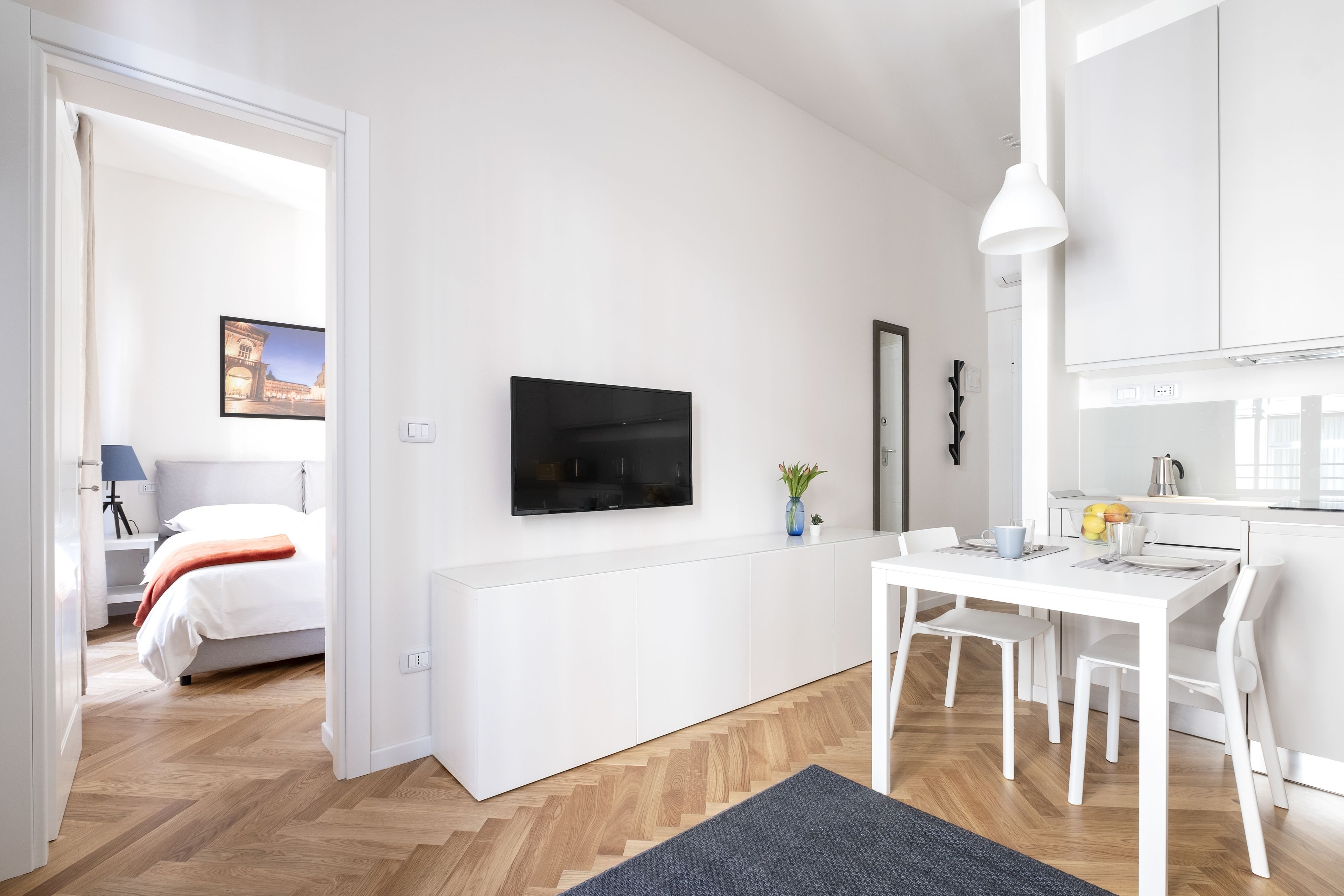 A modern, minimalist room with a bed, a TV mounted on the wall, a dining table with chairs, and a kitchen area.