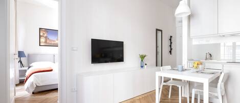 A modern, minimalist room with a bed, a TV mounted on the wall, a dining table with chairs, and a kitchen area.