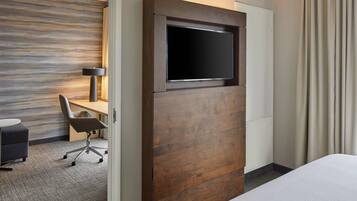 A hotel room with a bed, a desk with a chair, a television mounted on a wooden panel, and a lamp.