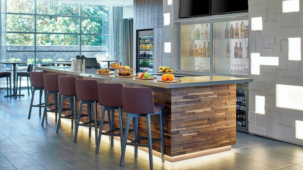 A modern bar area with a wooden counter, bar stools, and a large window offering a view of greenery. - Residence Inn by Marriott Albany Airport (Albany)