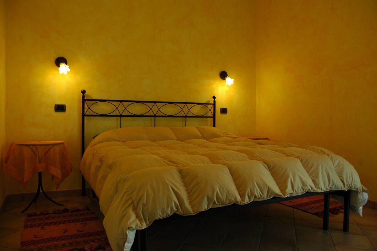 Ein Bett mit Kopfteil, zwei Wandleuchten, ein kleiner Tisch mit Stoffauflage und ein gemustertes Teppich.