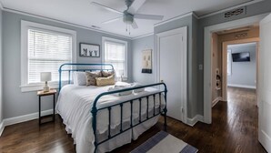 Un dormitorio con cama, ventilador de techo, mesita de noche, lámpara y televisor en la habitación contigua.