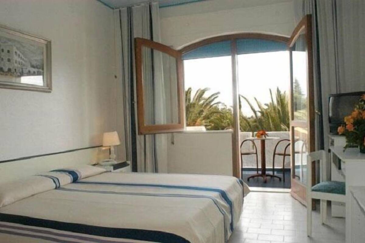 A hotel room with a bed, a desk, a chair, and a balcony with a view of greenery.