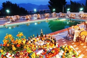 A poolside dining area with tables and chairs, a buffet with various food items, and a view of mountains in the background.