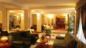 Un hall d’entrée spacieux, aménagé avec un canapé, des fauteuils et une table basse. On y trouve plusieurs lampes et des plantes en pot.