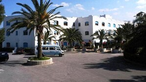Un bâtiment blanc avec de nombreuses fenêtres et un espace de stationnement où se trouve une voiture bleue et des palmiers.