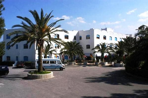 A white building with multiple windows and a parking area with a blue car and palm trees.