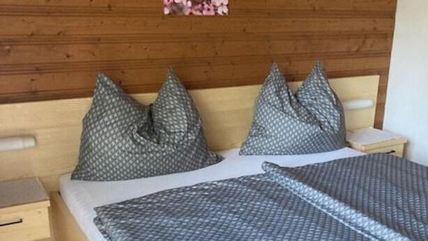 A bed with a patterned bedspread and two pillows, a wooden headboard, and a small bedside table with a drawer. - Double room 4 - Haus Bleckwand (Gschwendt)