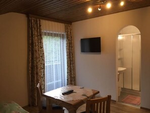 Une pièce avec un plafond en bois, une table avec une chaise, un téléviseur fixé au mur et une salle de bain visible à travers une arche.