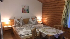A room with a bed, two bedside tables, a chair, a table with a tablecloth, and a wooden wall. - Double room 4 - Haus Bleckwand (Gschwendt)