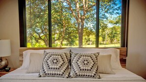 Un dormitorio con un ventanal grande, una cama con ropa de cama blanca y almohadas estampadas, y una mesita de noche con una lámpara y un reloj.