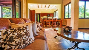 Un salón espacioso con un sofá de cuero, una zona de comedor con sillas de madera y grandes ventanales con vistas al verdor.