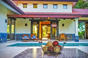 Zona de piscina con área de descanso cubierta, piscina con paredes de vidrio y una cesta decorativa con objetos.