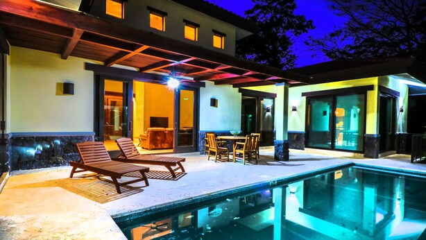Un área al lado de la piscina con sillones de descanso y una mesa de comedor, rodeada por una casa con ventanas grandes y un porche cubierto.