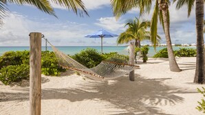 A hammock on a sandy beach with a view of the ocean and palm trees. - Altamer Resort (West End Village)