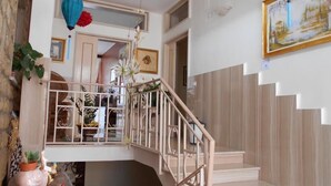 Una escalera con barandilla de madera y paredes blancas. Hay plantas en macetas en la rellano y un cuadro enmarcado en la pared.