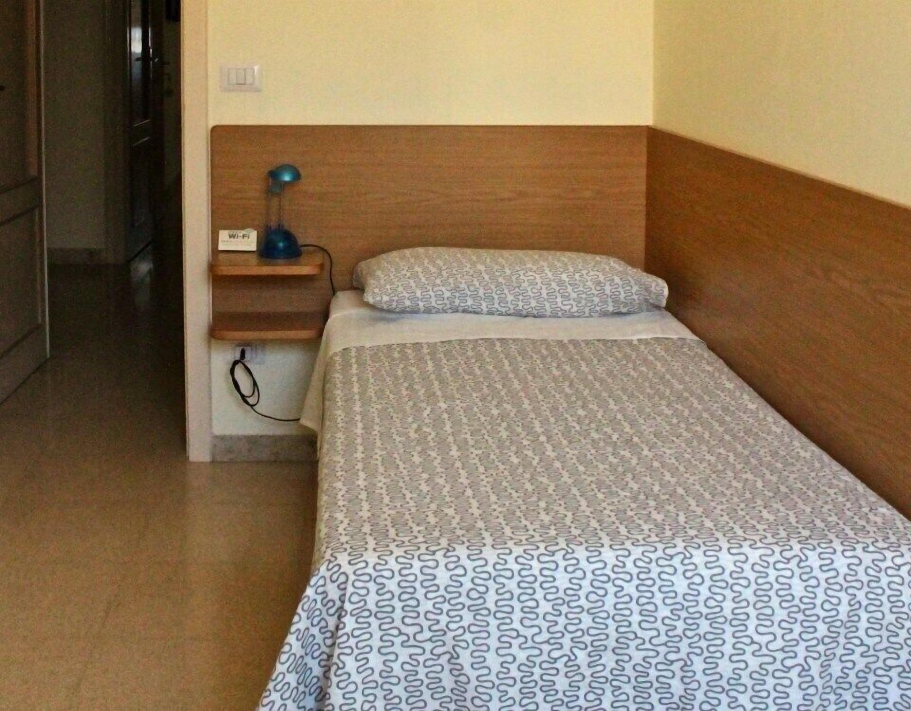 Una cama individual con una colcha estampada, cabecera de madera, lámpara de noche y una pequeña estantería con lámpara e enchufe.