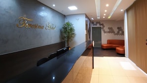 Un espace d’accueil d’hôtel comprenant un comptoir de réception, une plante en pot et un coin salon avec des chaises orange.