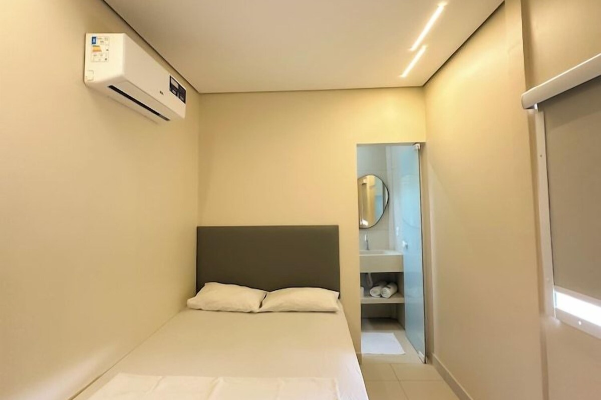 A narrow hotel room with a bed, a small bathroom with a mirror, and a wall-mounted air conditioning unit.