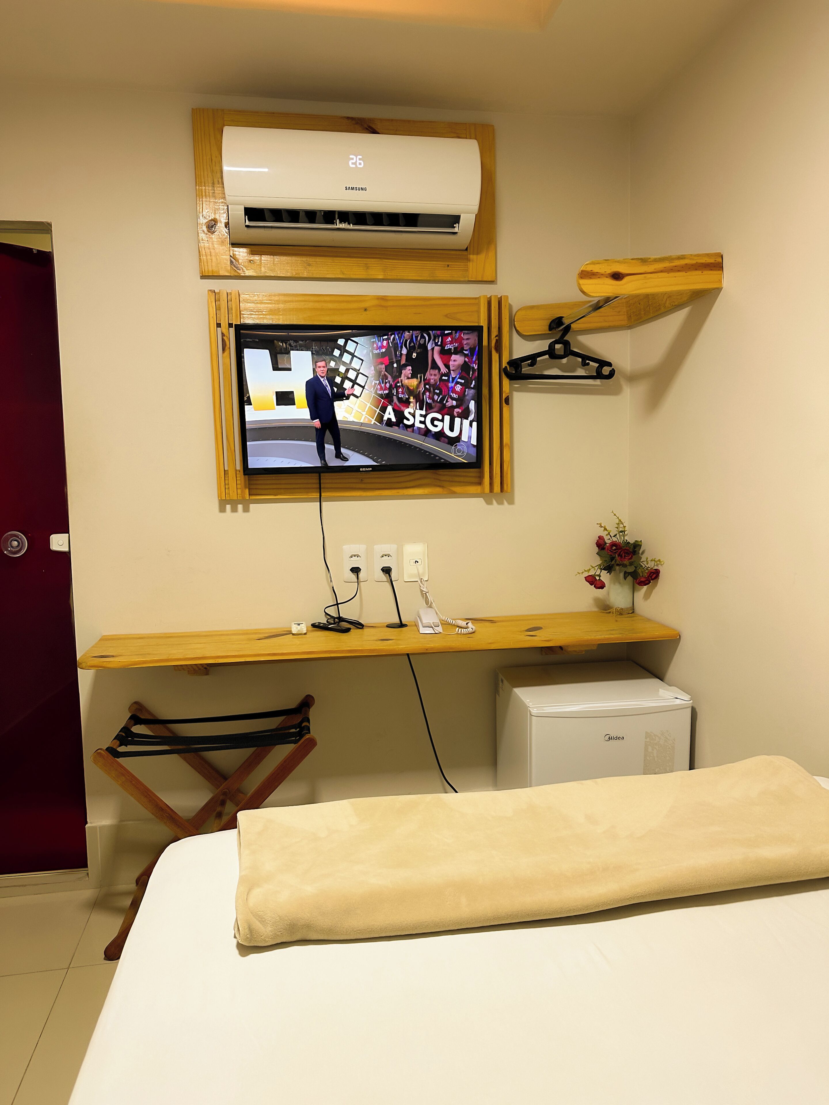 Una habitación de hotel con una cama, un escritorio de madera con un televisor, aire acondicionado y un refrigerador pequeño.