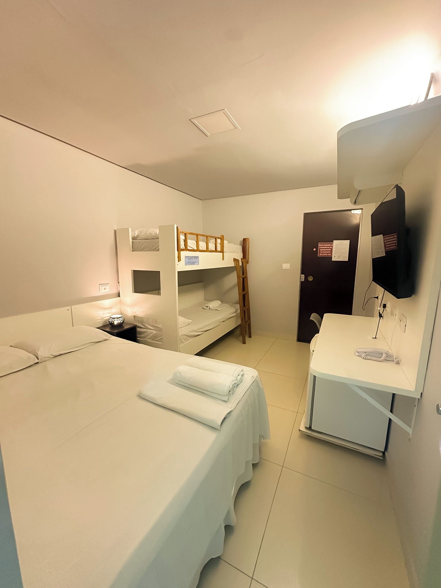 A single-bed room with a bunk bed, a desk, a chair, a mirror, and a door with a sign.