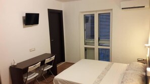A bedroom with a bed, a desk with chairs, a TV, and a window with blinds. - CasaMIA a Marina di Camerota (Camerota)
