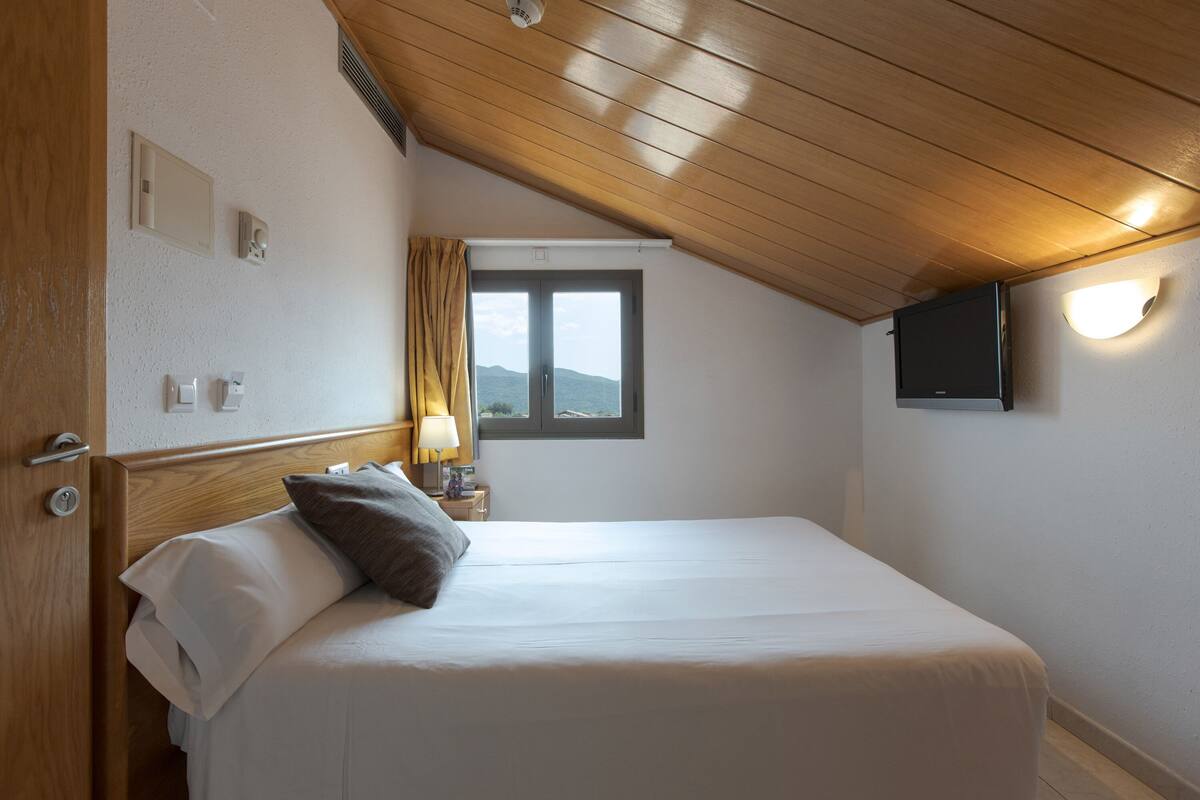 Un dormitorio con techo de madera, cama con ropa de cama blanca y marrón, mesita de noche con lámpara, ventana con vistas y televisor montado en la pared.