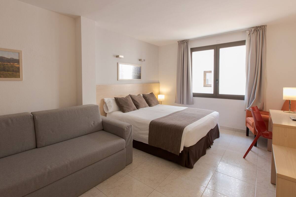 Habitación de hotel con cama, sofá gris, escritorio con silla y ventana con cortinas.