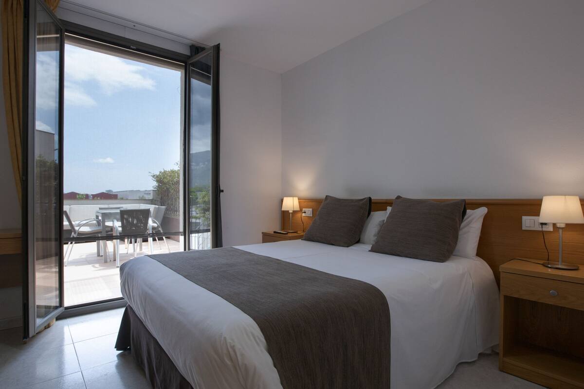 Habitación de hotel con una cama grande, dos mesitas de noche con lámparas, y un ventanal corredero que da a un espacio exterior con mesa y sillas.