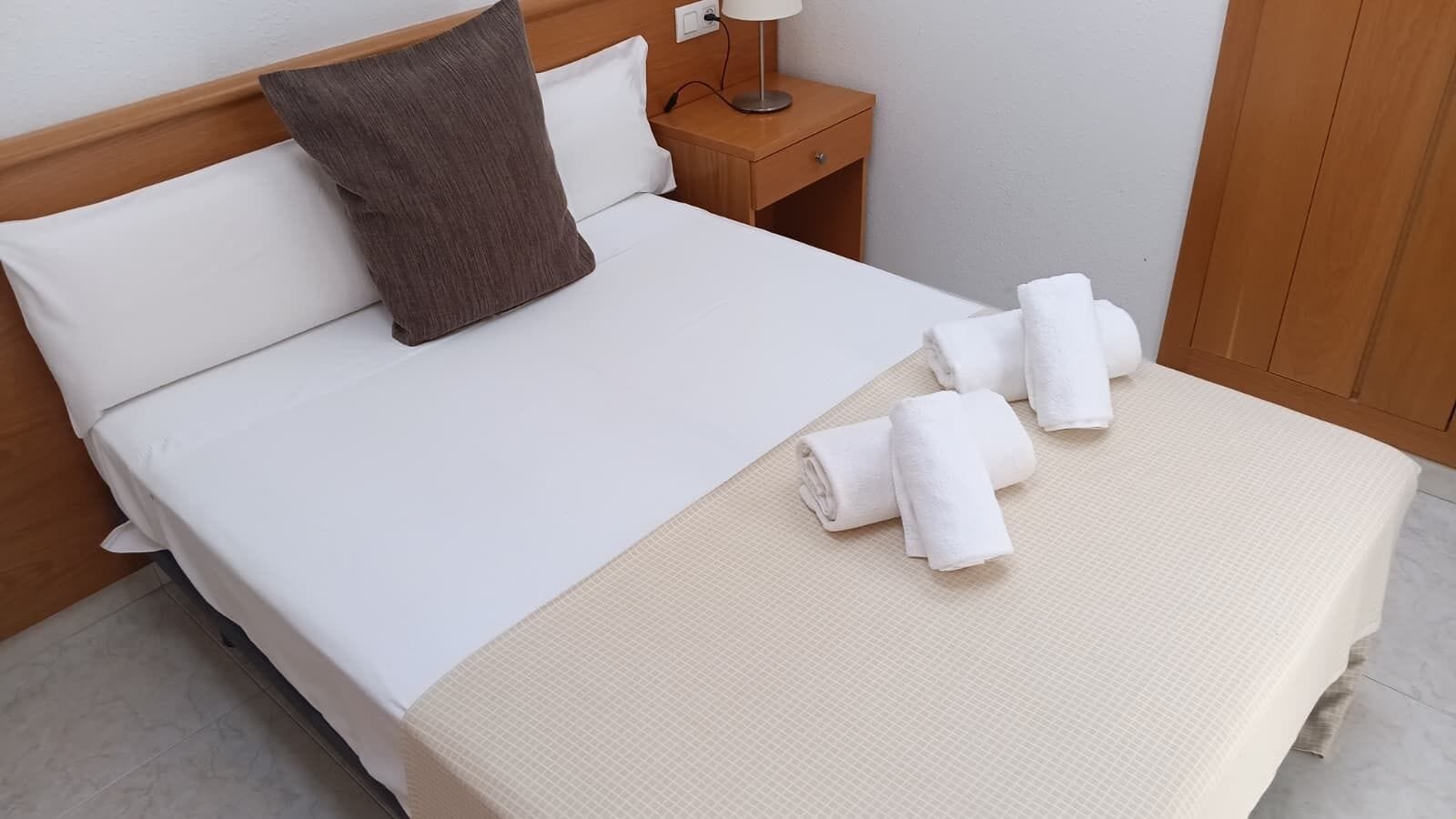 A neatly made bed with white linens, a brown pillow, and rolled towels on the bed.